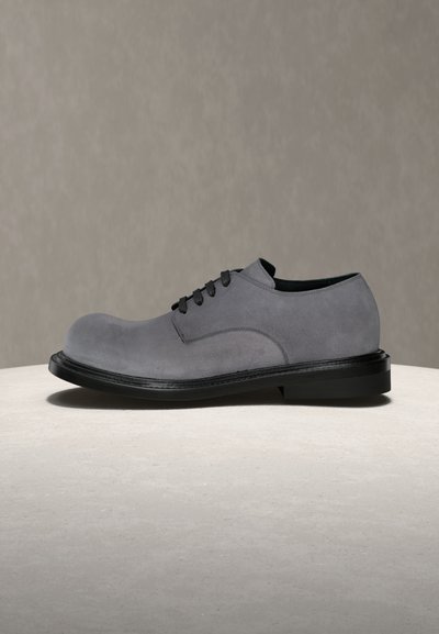 Chaussure derby en daim gris avec lacets noirs et semelle épaisse noire, présentée sur une surface en pierre claire contre un fond taupe atténué.