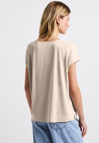 Beige top s krátkým rukávem a zaobleným výstřihem, hladkou texturou a volným střihem. Boční rozparky na lemu pro snadný pohyb.
