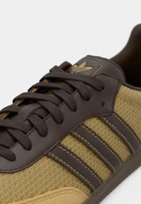 Bruna läderskor med tan-nät som har tre mörkbruna ränder, texturerad yta och en logopatch på tungan.