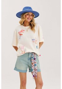 Krémové oversize tričko s farebnými potlačami v západnom štýle, skombinované s svetlomodrými denimovými šortkami a vzorovaným šálom na páse. Modrý klobúk.
