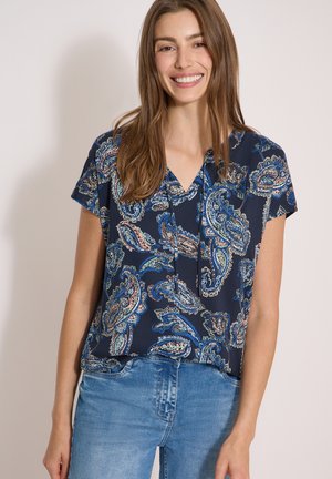 Junge Frau lächelnd, trägt eine blaue Paisley-Kurzarmbluse und hellblaue Jeans, steht vor einem schlichten hellen Hintergrund.