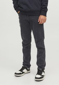 Dunkelgraue Jeans mit schlanker Passform, aus Denim gefertigt. Sie weisen dezente Ausbleichungen und eine einzelne Fronttasche auf. Kombiniert mit schwarz-weißen Turnschuhen.