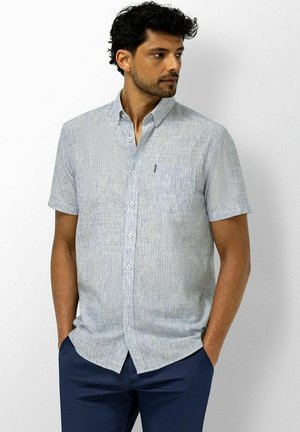 Man met kort krullend zwart haar, die een lichtgrijs kort mouwen overhemd met knopen en donkerblauwe broek draagt, staat met zijn handen in de zakken.