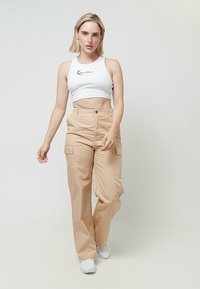 Tovorne hlače v bež tkanini, s poudarjenim visokim pasom, tovorni žepi in klasičnim zapiranjem na gumb. Kombinirane z belo kratko majico brez rokavov.