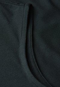 Tmavě zelená látka zblízka s žebrovanou texturou a jemným leskem. Čistý šev obrysy otvoru kapsy na levé straně.