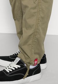 Khaki spodnie z elastycznymi mankietami i detalami wzdłuż boków, zestawione z czarnymi zamszowymi sneakersami z białymi podeszwami i sznurówkami, pokazującymi czerwoną metkę.