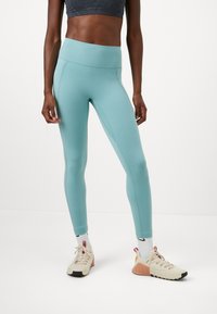Legginsy w kolorze niebiesko-zielonym z wysokim stanem, wykonane z elastycznego materiału. Charakteryzują się gładką teksturą i prostym wzorem, w połączeniu z beżowym obuwiem sportowym.