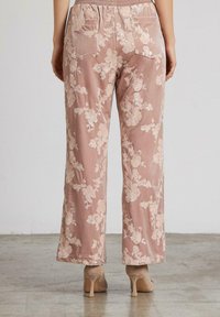 Donna che indossa pantaloni di velluto rosa con ricami floreali e tasche posteriori, abbinati a scarpe con tacco beige, in piedi su un pavimento in cemento.