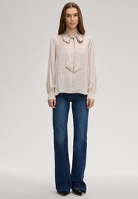 Lichtbeige zijden blouse met een striksluiting bij de hals, lange pofmouwen, gecombineerd met donkerblauwe uitlopende jeans en zwarte schoenen met hak.