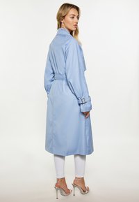 Trench coat bleu deschis cu talie cu curea, mâneci lungi și textură netedă, purtat peste pantaloni albi și sandale argintii cu curele.