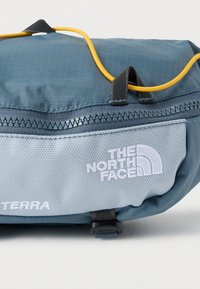 Μπλε και γκρι τσάντα North Face Terra με ορατό φερμουάρ, μαύρη θηλιά και κίτρινο κορδόνι σε λευκό φόντο.
