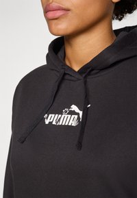Svart hettegenser laget av bomull, med en hvit Puma-logo med blomsterdesign aksenter. Inkluderer snorer i halsen.
