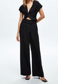 Černý crop top má design s twistingem uzlu, kombinovaný s vysokými širokými kalhotami. Oba kousky jsou vyrobeny z hladkého materiálu.