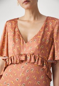 Femme portant une robe couleur rouille à col en V avec des motifs paisley orange et un volant froncé sur la poitrine et les manches.