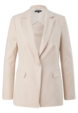 Femme portant un tailleur beige clair sur mesure avec un blazer à un bouton, un pantalon assorti, un haut rayé et des mocassins crème, debout devant un fond uni.