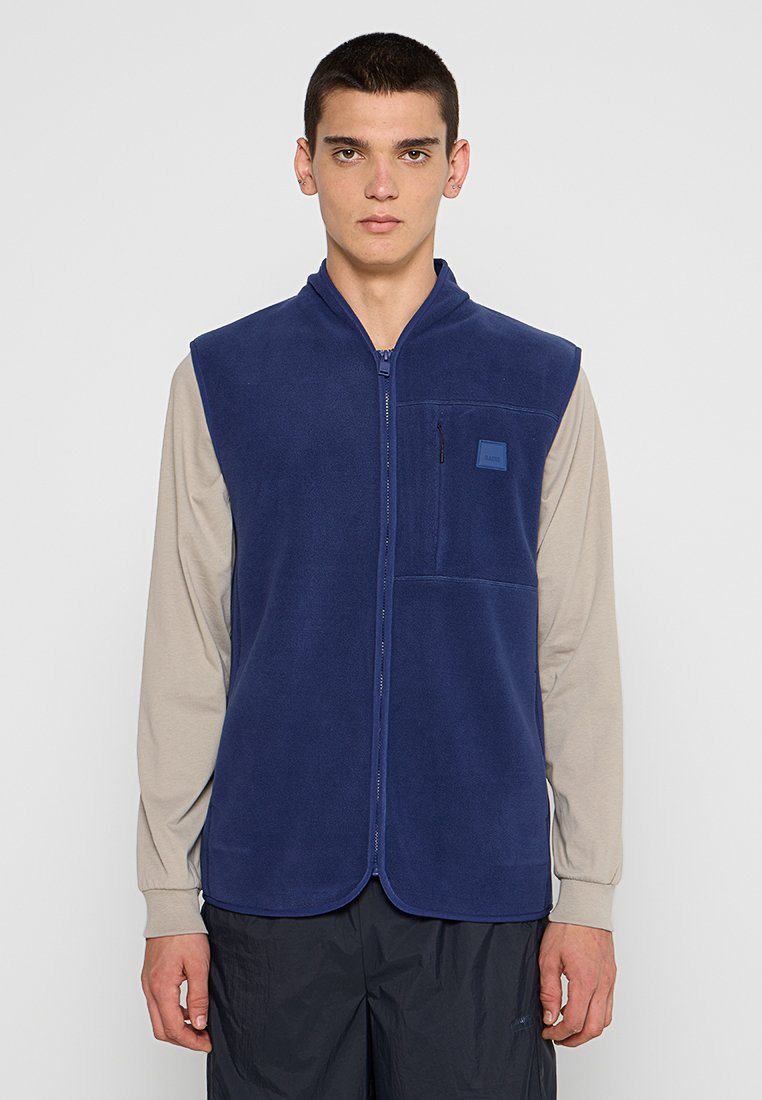 Gilet en polaire marine avec un col haut, une fermeture éclair frontale complète et une poche poitrine. Il présente une texture lisse et un design minimaliste.