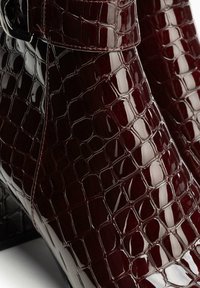 Burgundské lakované kozačky nad kotníky s krokodýlí texturou, s prvkem sponky a černým podpatkem. Hladký, lesklý povrch.