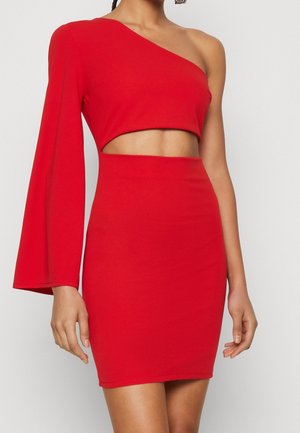Woman wearing a red one-shoulder dress with a cutout at the waist and a long sleeve on the left arm, standing against a plain background.