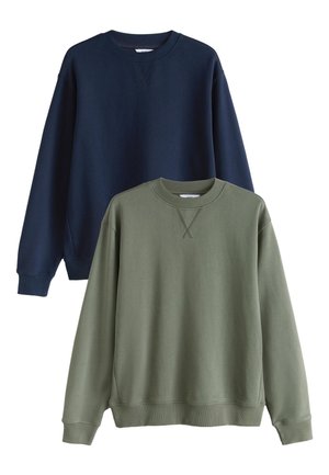 Junger Mann, der einen marineblauen Sweatshirt, dunkelgraue Jeans und schwarze Low-Top-Sneaker trägt, vor einem einfachen hellen Hintergrund.