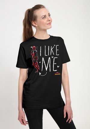 Kvinde iført sort T-shirt med Deadpool-grafik og teksten "I like me" i hvide bogstaver, stående mod en ensartet lys baggrund.