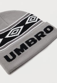 Gorro de malha cinza com padrão geométrico preto e branco e texto "UMBRO" na aba dobrada, apresentado plano sobre uma superfície branca.