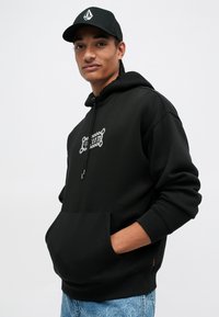 Un hoodie noir avec une poche avant, une capuche à cordon et des détails en logo argenté. Associé avec un jean bleu clair et une casquette noire avec un logo.