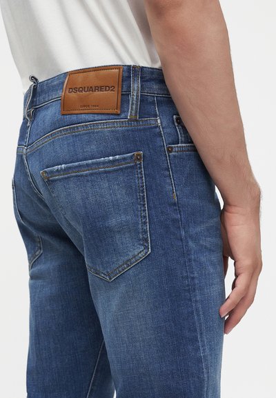 Gros plan d'une personne portant un jean en denim bleu avec une étiquette marron Dsquared2 à la taille et une chemise blanche rentrée.