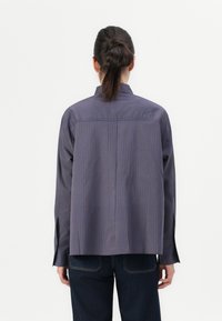 Une personne avec des cheveux foncés en queue de cheval portant une chemise à rayures bleu et marron à manches longues et un jean sombre, se tenant contre un fond blanc uni.