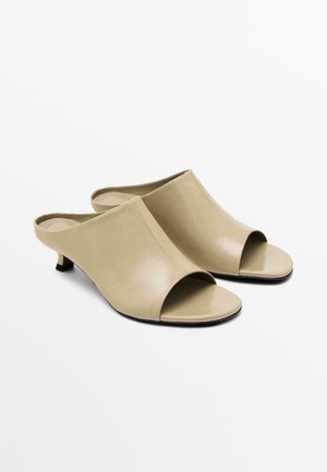 Mules ouvertes beiges en cuir lisse avec un petit talon, présentant un design minimaliste et des coutures le long des bords.