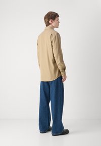 Cămașă button-up dintr-un material ribbed bej deschis, purtată cu pantaloni lejeri din denim albastru închis. Design simplu, cu o margine curbată și mansete.