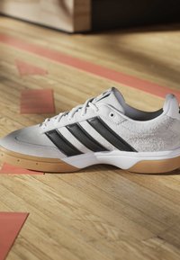 Weißer Sportschuh mit schwarzen Streifen und Gummisohle, der auf einem hölzernen Hallensportboden mit roten Markierungen liegt.