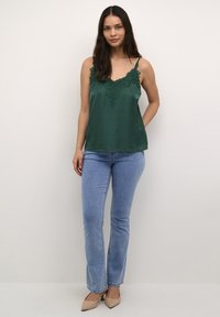 Satinova camisola v smaragdno zeleni barvi z čipkastim trimom na dekolteju, v kombinaciji s svetlo modrimi širokimi džinsami in bež čevlji s špicasto konico.