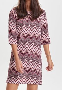Kobieta w sukience do kolan w różowo-białe-ciemnoczerwone wzory w zygzaki oraz rękawami o długości trzech ćwierci, stojąca na jednolitym tle.