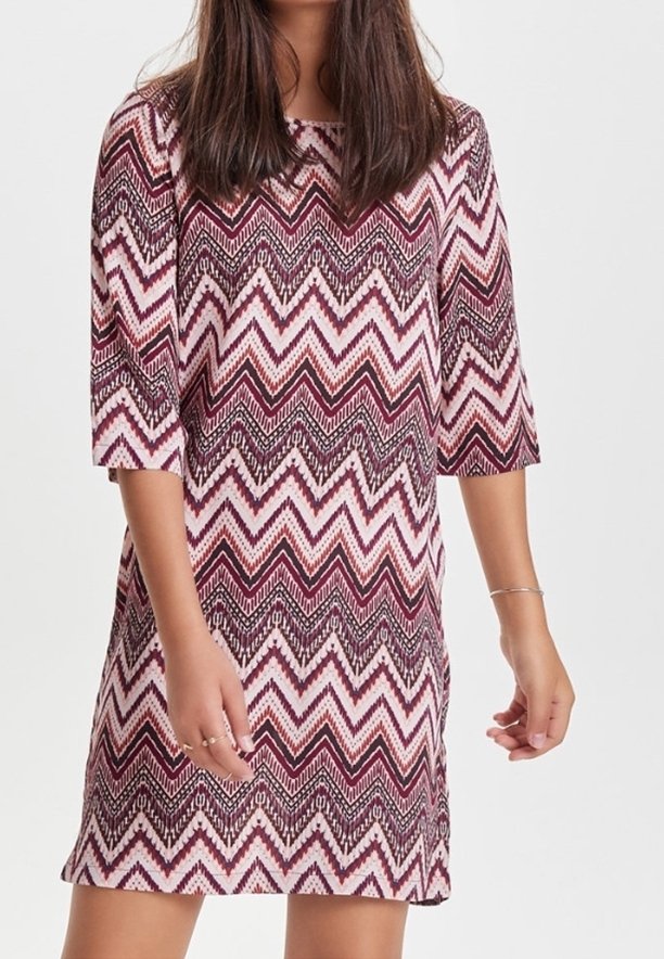 Kobieta w sukience do kolan w różowo-białe-ciemnoczerwone wzory w zygzaki oraz rękawami o długości trzech ćwierci, stojąca na jednolitym tle.