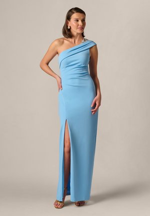 Kobieta w jasnoniebieskiej wieczorowej sukni na jedno ramię z rozcięciem na udzie, nosząca złote szpilki i duże kolczyki, stojąca na jednolitym tle.