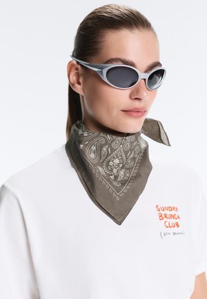 Donna con capelli pettinati all'indietro che indossa occhiali da sole grigi a forma di occhio di gatto, una bandana grigia fantasia e una camicia bianca con scritta arancione "Sunday Brunch Club."