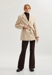Beige blazer met ceintuur, grote revers, knoopsluiting met één knoop en twee voorzakken, gecombineerd met bruine flared broeken en zwarte schoenen.