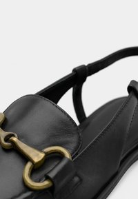 Μαύρο δερμάτινο παπούτσι με στρογγυλεμένη μύτη, λουράκι πίσω από το τακούνι και διακοσμητικό αγκράφα σε μπρούτζινο χρώμα. Ομαλή υφή με διακριτικές λεπτομέρειες ραφής.