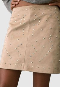 Minifalda de ante sintético beige con tachuelas metálicas dispersas en varios tamaños, que crean una superficie texturizada con un brillo sutil.