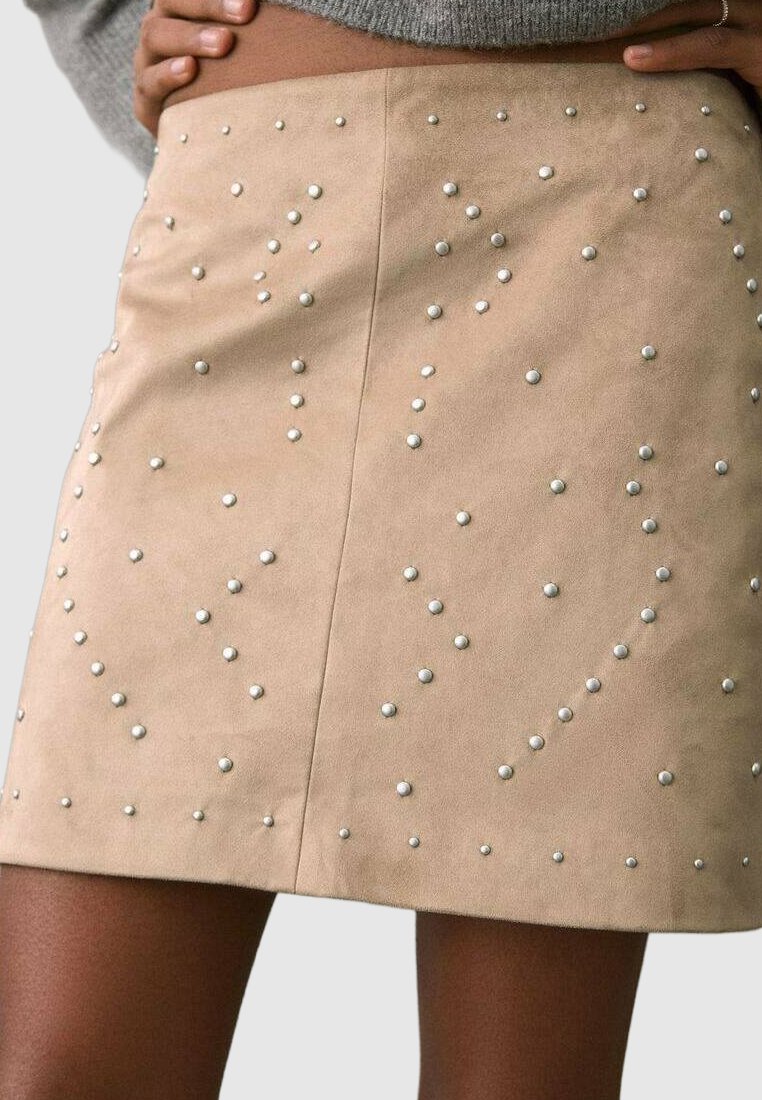 Minifalda de ante sintético beige con tachuelas metálicas dispersas en varios tamaños, que crean una superficie texturizada con un brillo sutil.