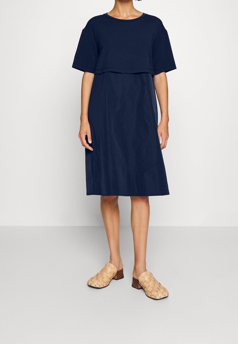 Vrouw draagt een marineblauwe midijurk met korte mouwen en een gelaagde bovenkant, gecombineerd met beige gevlochten leren mules met lage hakken.