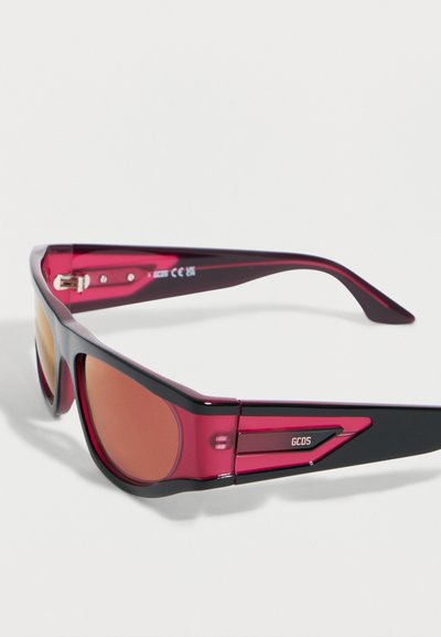 Ορθογώνια γυαλιά ηλίου με κόκκινους και μαύρους γυαλιστερούς σκελετούς και φακούς με κεχριμπαρένιο χρώμα, με λογότυπο GCDS στο πλάι.