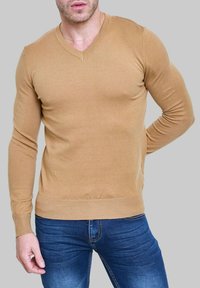 Pull beige à col en V et manches longues, en tissu tricoté doux, avec un design ajusté et des poignets et un ourlet côtelés, associé à un jean bleu.