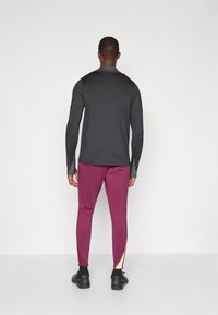 Camisola desportiva de mangas longas preta com detalhes cinzentos, combinada com calças justas bordeaux com detalhes em painel lateral e sapatilhas desportivas pretas.