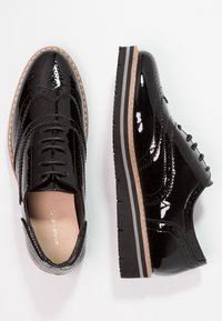 Des chaussures oxford en cuir verni noir présentent des détails de brogue, un design à lacets et une semelle texturée contrastante avec une bande de couleur claire.