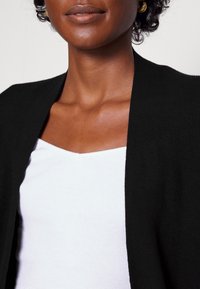 Cardigan noir en maille avec un devant ouvert, porté sur un haut blanc côtelé, présentant une texture lisse et un design épuré.