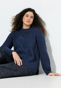 Marineblauer Strickpullover mit Rundhalsausschnitt und gesticktem Ampersand-Design. Kombiniert mit gemusterten dunkelblauen Hosen mit Leopardenmuster.