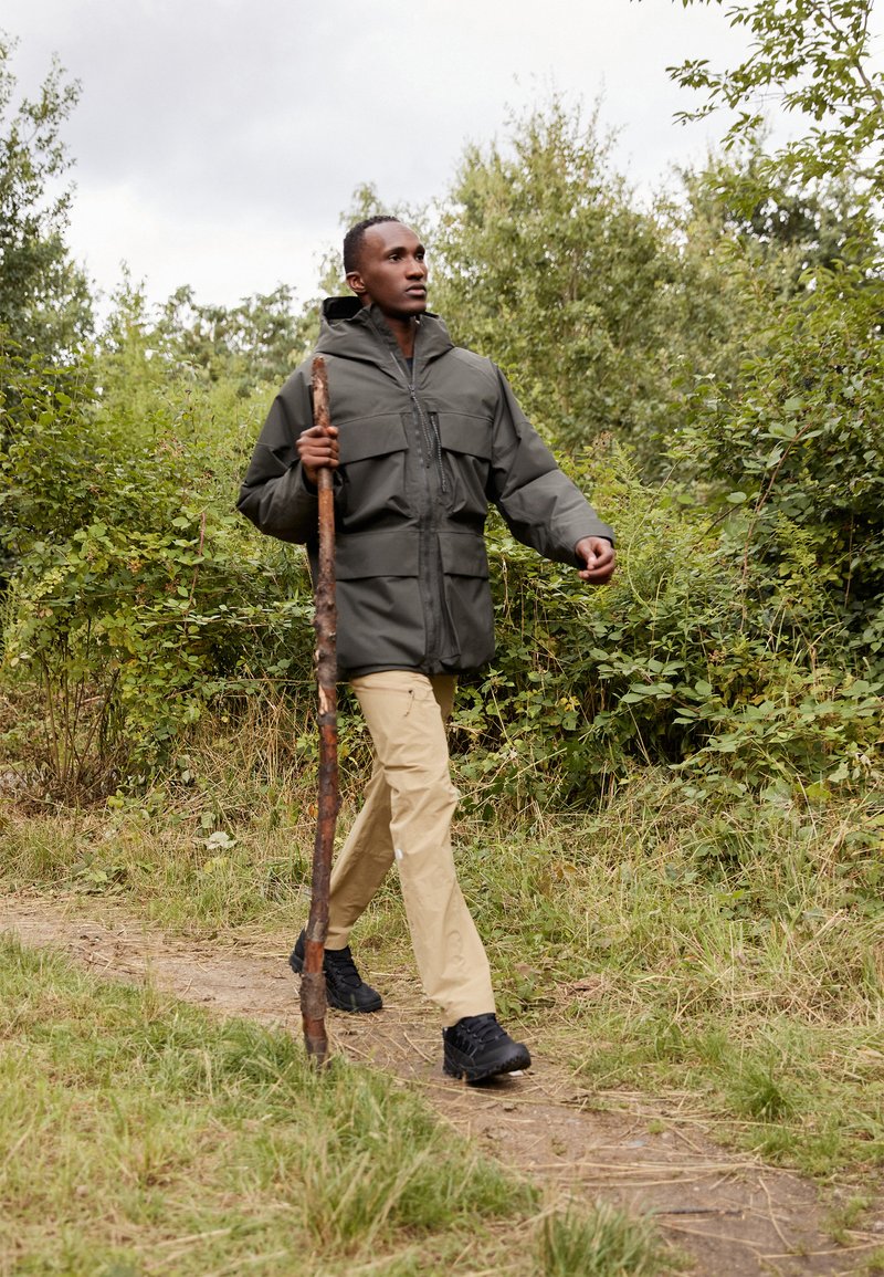 Chaqueta exterior en verde oscuro, pantalones beige y zapatos negros. El modelo sostiene un bastón de madera, caminando por un sendero natural rodeado de vegetación.