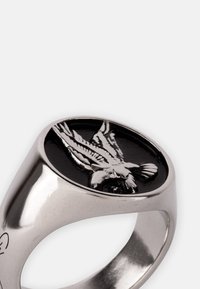 Anillo de plata con un centro ovalado negro y un diseño de águila plateada en relieve. Superficie suave, acabado pulido y forma moderna.