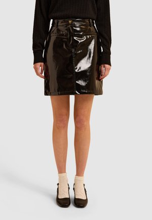 Jupe mini en cuir verni noir avec une fermeture éclair à l'avant, des passants de ceinture et des poches latérales. Portée avec des chaussettes crémeuses texturées et des chaussures sombres.